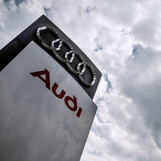 Dunkle Wolken über einen Audi-Logo (Symbolbild). Gerüchte um Kapazitätskürzungen und Sparpläne sorgen für Unruhe bei Audi. Besonders das Werk Neckarsulm ist in den Fokus gerückt. 
