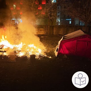Ein Feuer auf einem Stück Wiese. Daneben steht ein Zelt. Eine Dinge sind verbrannt. Auch Kälte·iglus für Wohnungs·lose sind verbrannt. Im Hintergrund sind Häuser und eine Mauer. Rechts das SWR Logo für Leichte Sprache.