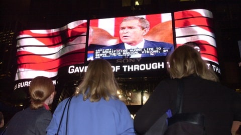 Menschen betrachten auf einer Leinwand eine TV-Stellungnahme von Präsident George W. Bush