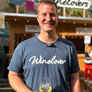 Moritz Ocker ist der erste Weinprinz Württembergs. Er steht vor einem Weinstand und hält ein Glas Wein in der Hand.