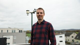 Benjamin ist Sturmjäger. Er steht draußen und im Hintergrund sind graue Wolken zu sehen. Er trägt ein rot-schwarz-kariertes Hemd und lacht in die Kamera.