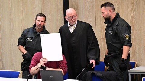 Der Angeklagte in einem Mordprozess nach einer Todesfahrt von Mannheim sitzt mit einem Aktendeckel vor seinem Gesicht im Gerichtssaal, rechts neben ihm steht sein Verteidiger. 