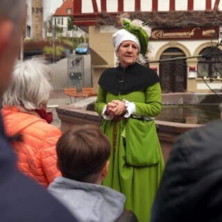 Eine Stadtführerin in grünen Mittelalter-Gewändern steht vor einem Brunnen und redet zu einer Gruppe von Leuten.