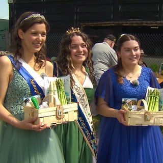 Drei junge Frauen: Emilia, amtierende Schwetzinger Spargelkönigin, Franziska, Spargelkönigin von Schrobenhausen und Marlen, zukünftige Schwetzinger Spargelprinzessin. Alle tragen schicke Kleider und Emilia und Marlen halten Körbe in den Händen, in denen Spargel ist.  