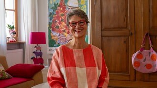 Ältere Frau mit grauen Haaren und roter Brille sitzt in ihrem Wohnzimmer und lächelt in die Kamera.