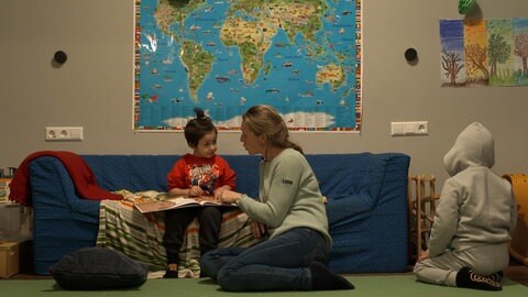 Ein Kind sitzt in der Spielstube Stuttgart Weilimdorf auf einem Sofa und schaut sich gemeinsam mit einer Betreuerin ein Buch an.