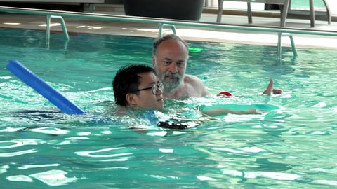 Chinesischer Student schwimmt mit einer Schwimmnudel und mit Hilfe eines Schwimmlehrers