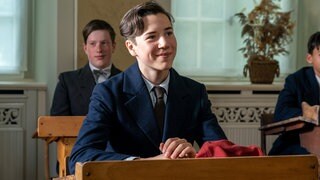Herbert (Arthur Gropp) sitzt an seinem Platz im Klassenzimmer und lächelt.