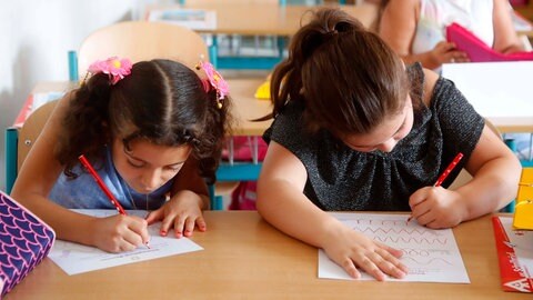 Zwei Mädchen schreiben, eines mit der linken Hand, das andere mit der rechten. Die richtige Schreibhaltung wird Kindern, die mit links schreiben, nur selten richtig beigebracht - gezielte Unterstützung für Linkshänder und ihre Besonderheiten bleibt meist aus.