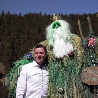 Ben steht in seinem grünen Mummelsee-Kostüm und mit einer weißen Langhaar- und Bartperücke am Mummelsee. In der rechten Hand hält er einen goldenen Dreizack, den linken Arm hat er um Hotelmanager Karl-Heinz Müller gelegt.
