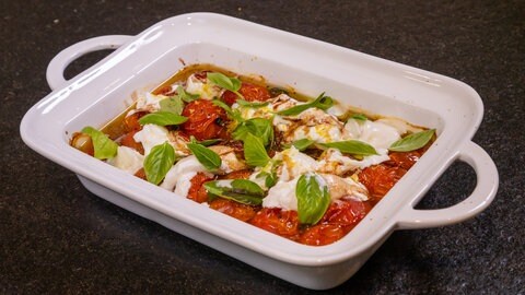 Das fertige Ofengericht in einer Auflaufform. Die gebackenen Tomaten sind mit zerstückelter Burrata und mit Basilikum bedeckt.
