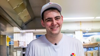 Junger Mann in Bäckerkleidung steht in einer Bäckerei und lächelt in die Kamera