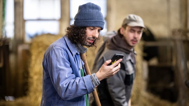 Albert (Alessio Hirschkorn) und Sebastian (Dominik Stricker) arbeiten im Stall, Albert blickt auf sein Handy