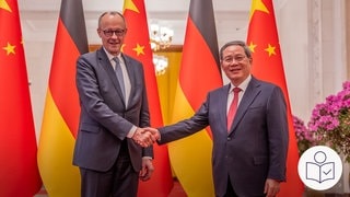Bundeskanzler Merz und Chinas Regierungs·chef Li stehen nebeneinander vor großen Fahnen. Rechts das SWR Logo für Leichte Sprache. 