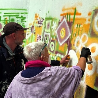 Graffiti-Workshop in Mannheim: Ein Mann mit Kappe und eine Frau mit grauem Haar besprühen Wand.