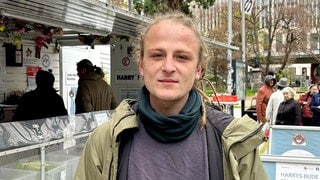 Lukas steht vor „Harrys Bude” in der Tübinger Straße in Stuttgart. Im Hintergrund ist der Wagen von „Harrys Bude” zu sehen. Und Menschen, die sich für Lebensmittel anstellen. Lukas schaut in die Kamera. 