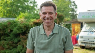 Mann mit braunen Haaren trägt ein grünes Tshirt und lächelt in die Kamera. Im Hintergrund ist ein Wohnmobil zu sehen.