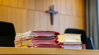 Akten liegen bei einem Prozess im Gerichtssaal. Im Hintergrund ist ein Kreuz an der Wand zu sehen.