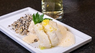 Kabeljau aus dem Ofen mit Sauce und Reis: Ein helles Fischfilet liegt auf Reis mit Sesamkörnern und ist mit einer hellen Sauce übergossen. Das Ganze ist schön auf einem weißen rechteckigen Teller angerichtet.