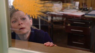 Frau Heilert duckt sich hinter einem Fenster, auf dem ein gelber Paintball-Fleck zu sehen ist