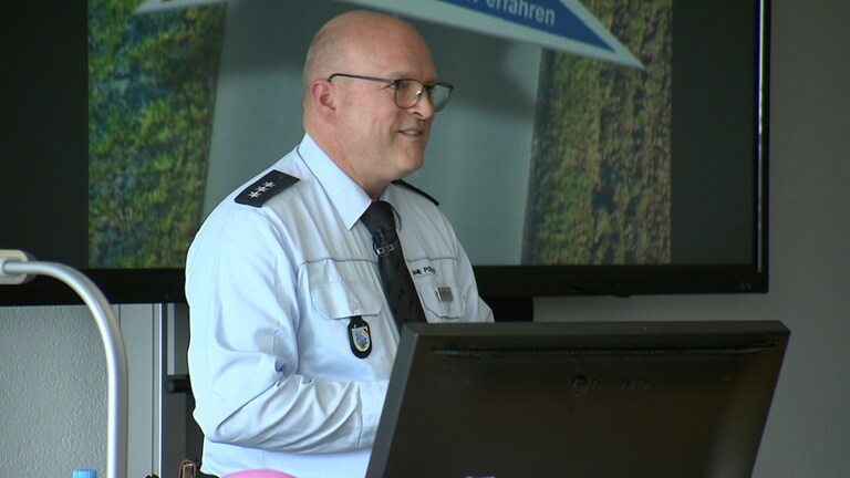 Ein Mann in Polizei-Uniform und Brille steht vor einer Klasse am Pult. Vor ihm befindet sich ein Bildschirm.