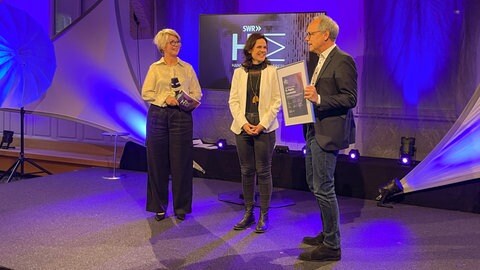 Regina Cazzamatta (Mitte) bei der Preisübergabe zusammen mit Kai Gniffke (rechts) und Moderatorin Stefanie Germann (links). Regina Cazzamatta ist Preisträgerin des Hans Bausch Mediapreis 2025.