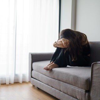 Eine junge Frau sitzt trauernd alleine auf dem Sofa und verbirgt ihr Gesicht | Trauer und finanzielle Sorgen: Jung verwitwet: Wenn der Partner zu früh stirbt