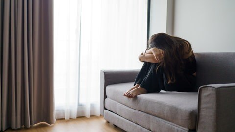 Eine junge Frau sitzt trauernd alleine auf dem Sofa und verbirgt ihr Gesicht | Trauer und finanzielle Sorgen: Jung verwitwet: Wenn der Partner zu früh stirbt