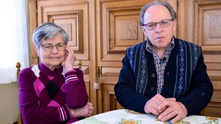 Karl Heinz und Magdalena sitzen an einem Tisch in ihrem Wohnzimmer in Hechingen. Beide tragen Brillen, Magdalena trägt einen lila Pullover und Karl Heinz eine dunkelblaue Strickweste. Im Hintergrund sind holzgetäfelte Türen zu sehen.