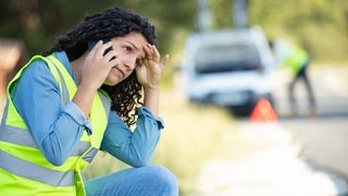 Eine Frau sitzt in Warnweste vor ihrem Auto und ruft jemanden an. Durch reflektierende Farben sollen Warnwesten in Notsituationen Sicherheit geben. Doch was taugt das leuchtende Material, wenn es darauf ankommt?   