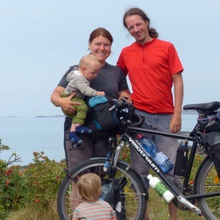 Dina und Rafael Streib stehen hinter einem schwarzen Fahrrad mit einem Kinderwagen-Anhänger. Auf ihrem Arm und vor dem Fahrrad sind zwei ihrer Kinder. Im Hintergrund erkennt man eine Küste.