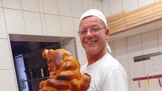 Ein Mann in Bäckerkleidung hält einen Hefezopf. Er lächelt.
