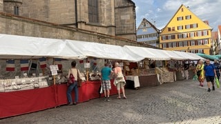 Eine Händlerin steht hinter einem bunt gefüllten Marktstand. Vor ihr liegen unter anderem Knoblauch, Gemüse und Blumen, über dem Stand hängen französische Fähnchen.