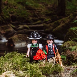 Die Playmobil-Figuren "Schwarzwald Marie" und Hannes in traditioneller Kleidung mit Bollenhut und Trompete in der Hand stehen im Wald.