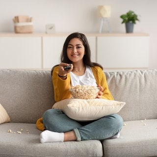 Bereit für den Serien-Marathon: Eine Frau mit Popcorn und Fernbedienung auf dem Sofa