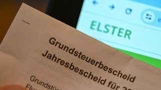 Ein Grundsteuerbescheid wird vor die Internetseite der Steuerplattform Elster gehalten.