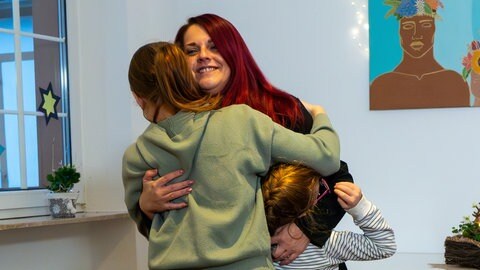 Eine Frau mit roten Haaren umarmt ihre beiden Kinder und lächelt über das ganze Gesicht.