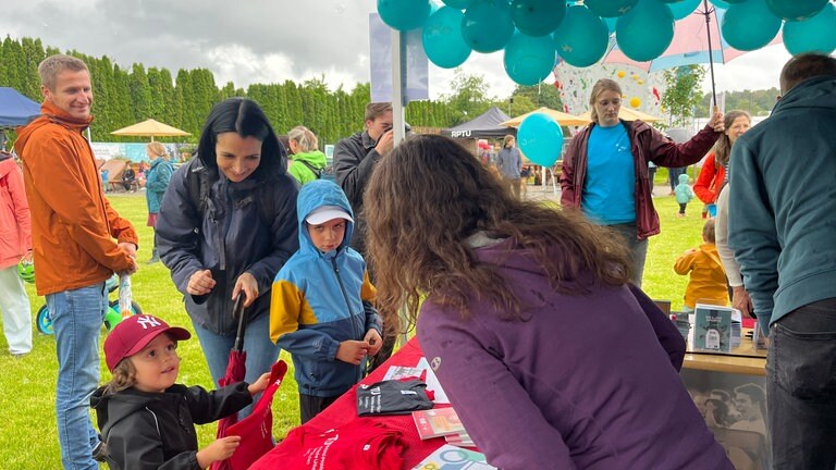 Ein Kind steht mit einem Shirt an einem Stand des Unisport: Der Unisport der Rheinland-Pfälzischen Technischen Universität Kaiserslautern-Landau (RPTU) hat auf dem Campus einen Abenteuerspielplatz eröffnet. Das Kitz-Land können Kinder mit einer Unisport-Karte besuchen.