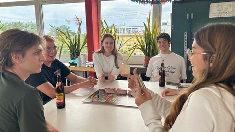 Sechs Studentinnen und Studenten bei ihrem wöchentlichen Spieleabend in Karlsruhe. Sie spielen ein Brettspiel auf einem Tisch.