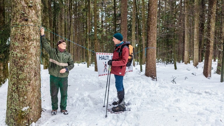 Beas Feriengast, Herr Balser, steht in Schneeschuhausrüstung im Wald bei Karl, der Verbotshinweise aufhängt