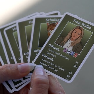 Eine Hand mit weißen Gelnägeln hält einige grüne Sammelkarten in der Hand. Auf der vordersten Karte steht der Name "Frau Walko", darunter eine Zeichnung von einer Frau mit blonden, langen Haaren.