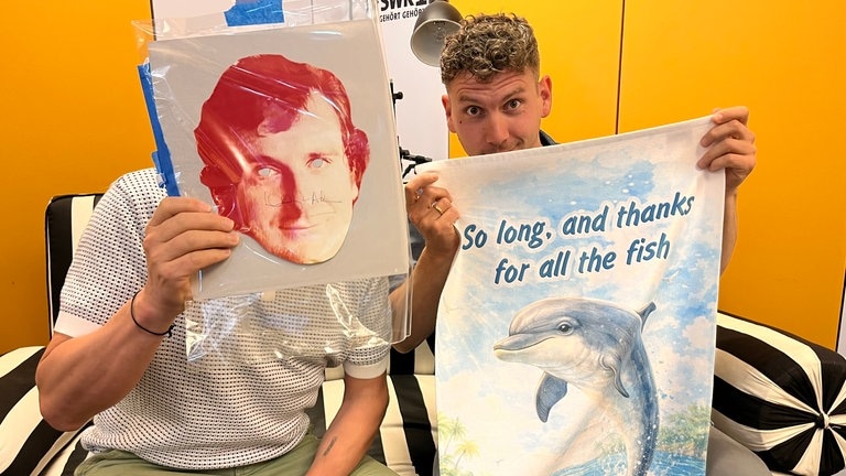Olly Krimmel zeigt eine handsignierte Maske von Douglas Adams, SWR1 Moderator Julian Hammerstein ein Handtuch mit dem Spruch "No panic. Thanks for all The Fish".