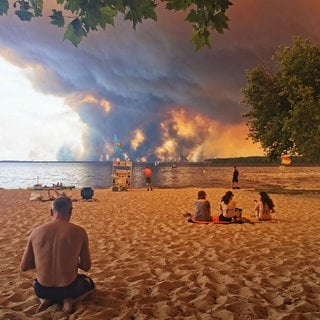 Strandbesuchende sitzen am Strand von Moutchic in Lacanau. Im Hintergrund sind Rauchwolken von den Waldbränden.