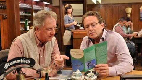 Schorsch und Bernhard sitzen am Stammtisch und schauen sich einen Prospekt an, Eva im Hintergrund am Tresen