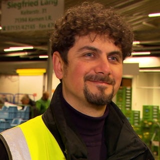 Christian Polder steht mit einer gelben Warnweste vor einer hell ausgeleuchteten Lagerhalle. Er hat lockige, braune Haare und einen Goatee. Er lächelt in die Kamera.