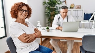 Frau sitzt zur Beratung in der Sprechstunde eines Arztes und lächelt: Über "die Wechseljahre" wissen Frauen oft wenig. Etwa dass das Östrogen schon ab Anfang 40 zu sinken beginnt. Und dass die Hormonersatztherapie gut wirkt und weitgehend sicher ist.
