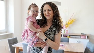Laura hält ihre 2-jährige Tochter Elisa auf dem Arm. Elisa hat das Angelman-Syndrom und trägt eine Brille, Laura hat braune Locken und trägt ein Sommerkleid. Beide lachen in die Kamera.
