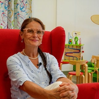 Eine Frau mit dunklen Haaren, Zopf und Brille sitzt auf einem roten Sessel.