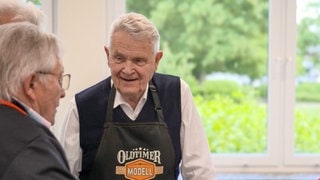 Dietmar ist im Gespräch mit zwei anderen Teilnehmern des Kochkurses für Herren über 70. Er hat weiße Haare, trägt einen dunklen Pullunder und eine schwarze Kochschürze, auf der „Oldtimer Modell“ steht. 