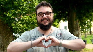 Mann mit Brille lacht in die Kamera und formt ein Herz mit seinen Händen.